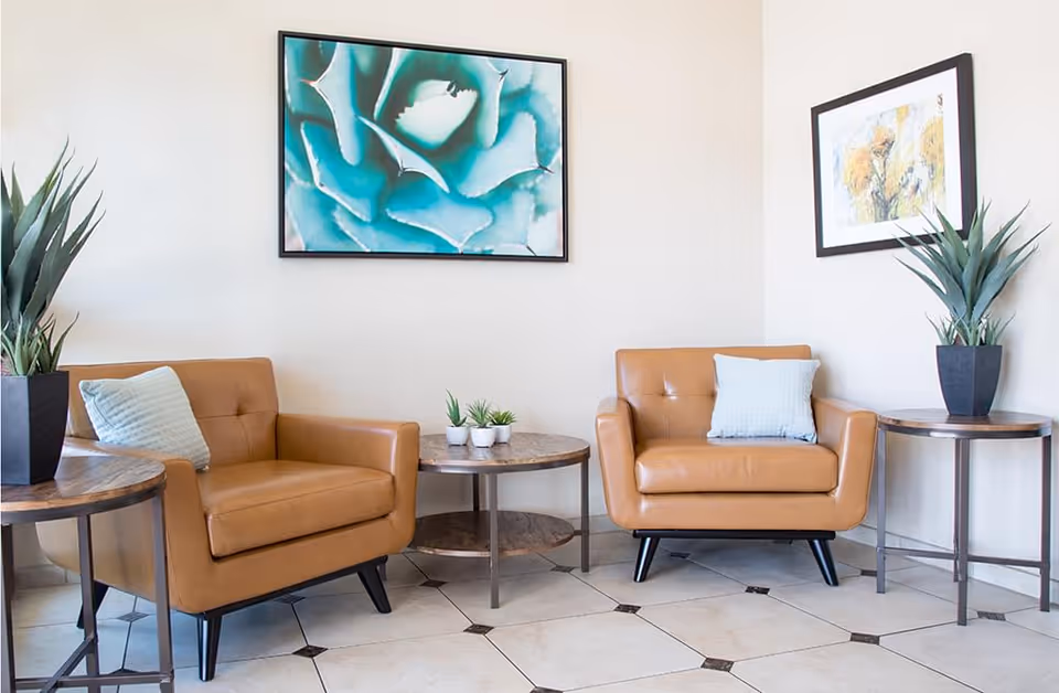 Two tan leather armchairs with side tables, potted plants and wall art in a bright sitting area.