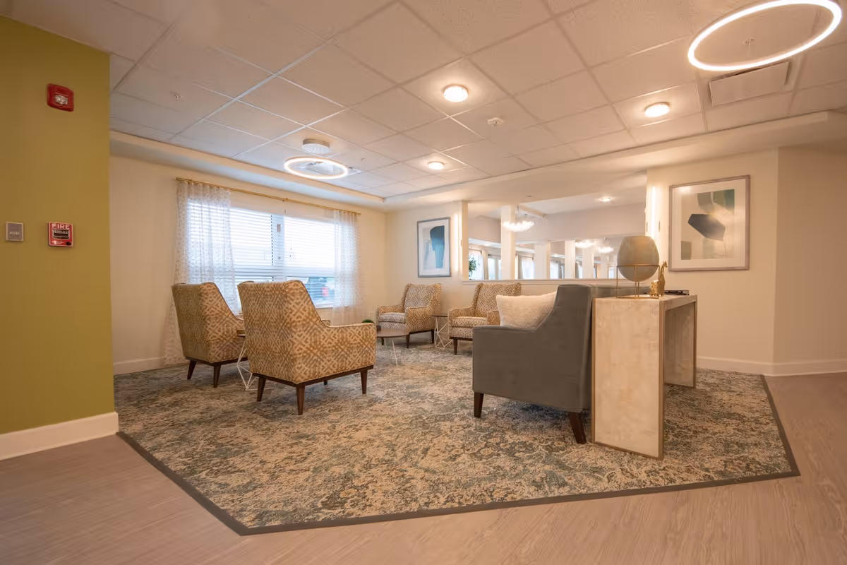 A bright communal lounge with patterned armchairs and a sofa arranged on a decorative rug beneath modern ceiling lights.