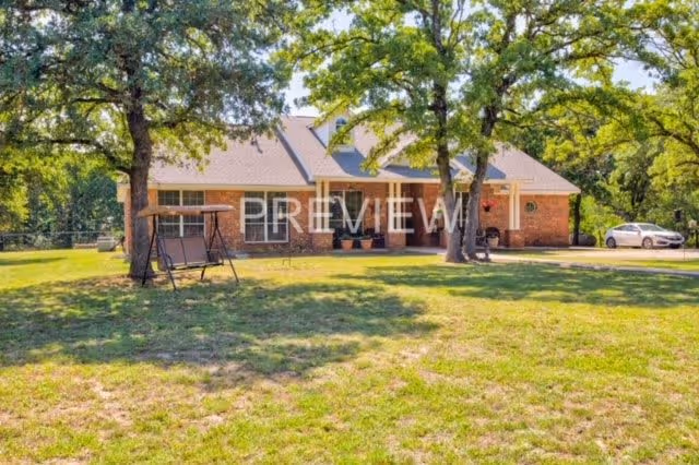 A single-story brick house with a gray roof surrounded by trees and a grassy yard. There is a swing set under one of the trees and a white car parked near the house. The scene is bright and sunny.