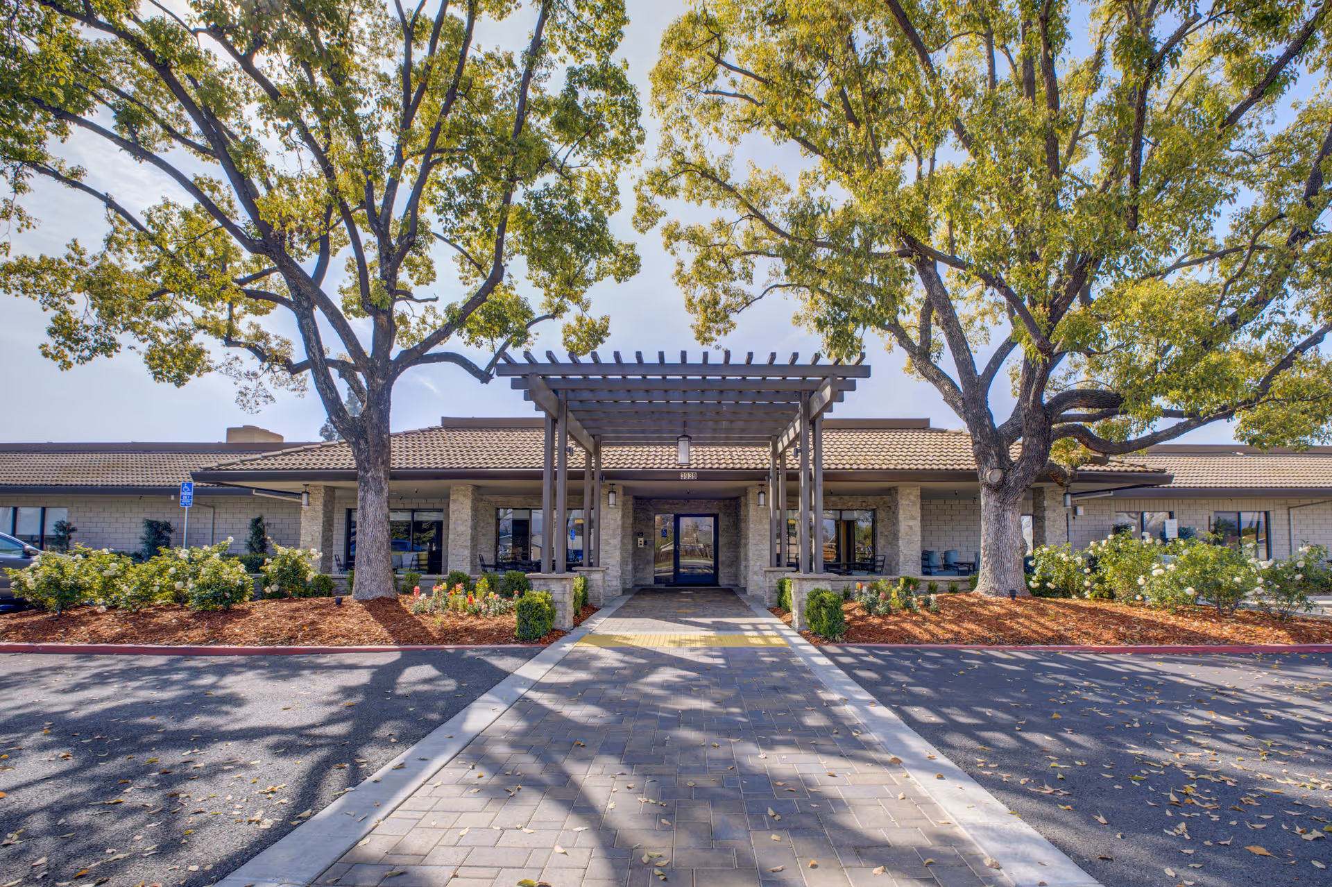 Front exterior view of Park Visalia Assisted Living & Memory Care building with a paved walkway leading to the entrance, flanked by two large trees and landscaped bushes.