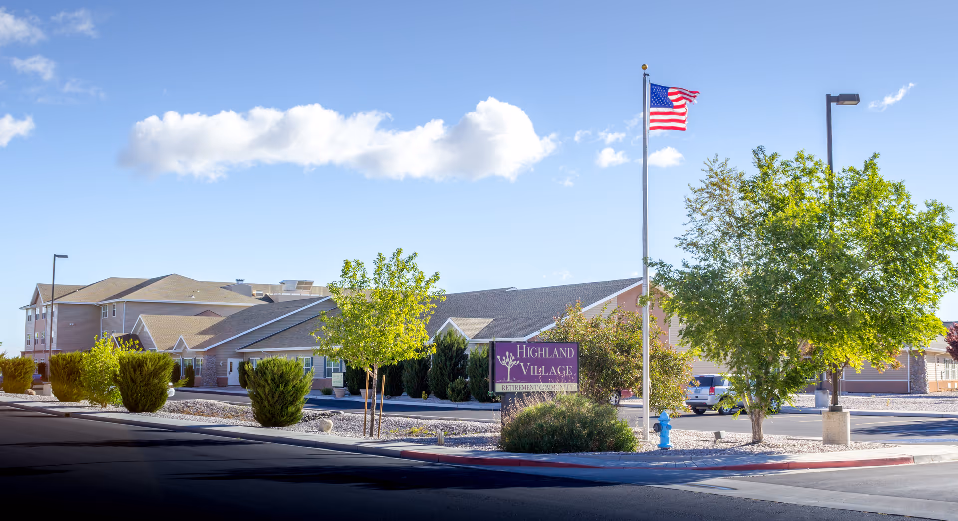 Front exterior of the Highland Village senior living building with its sign, landscaping, and an American flag flying on a flagpole.