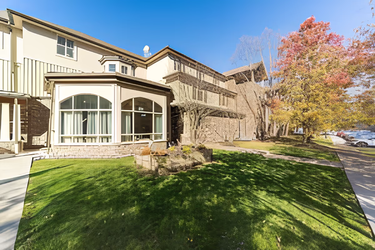 Exterior view of a senior living facility building with large windows, a well-maintained green lawn, and trees with autumn-colored leaves under a clear blue sky.