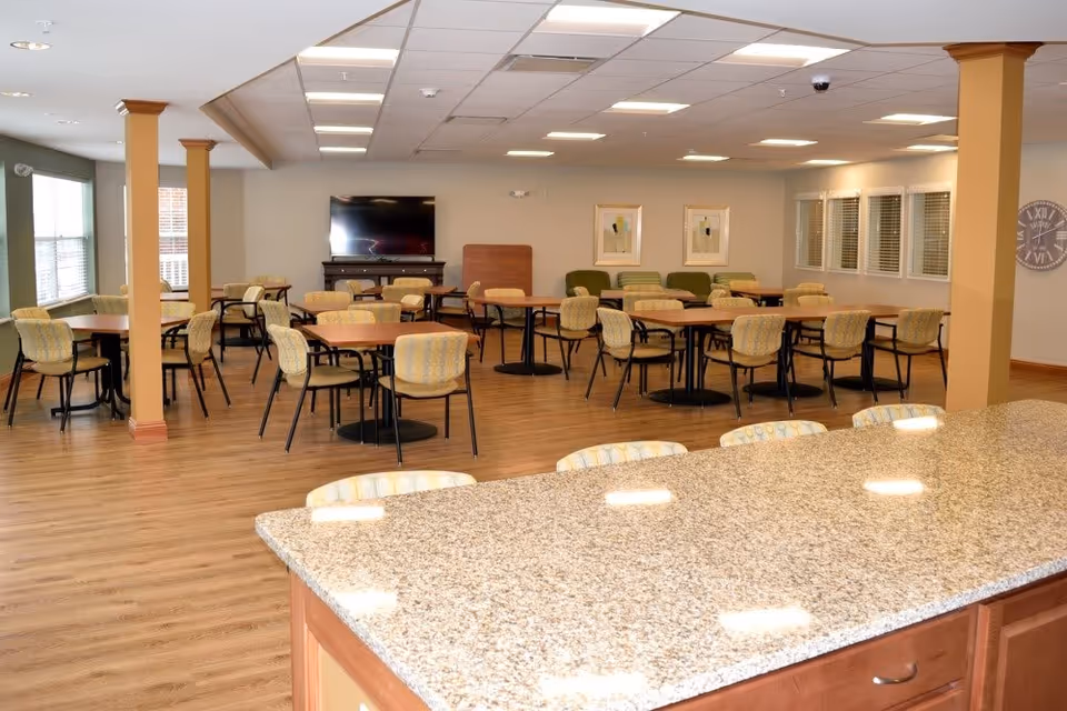 A spacious dining room with multiple round tables and chairs arranged neatly. The room has wooden flooring, beige walls, and a large flat-screen TV mounted on the far wall. There are windows with blinds on the right side and two framed artworks on the wall. In the foreground, there is a granite countertop with several chairs lined up along it.