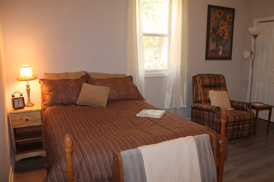 A cozy bedroom with a wooden bed covered in a brown striped bedspread and multiple pillows. A small wooden nightstand with a lamp and an alarm clock is on the left side of the bed. A book lies on the bed. To the right, there is a striped armchair with a cushion, a small wooden side table, a floor lamp, and a framed floral painting on the wall. A window with white curtains lets in natural light.