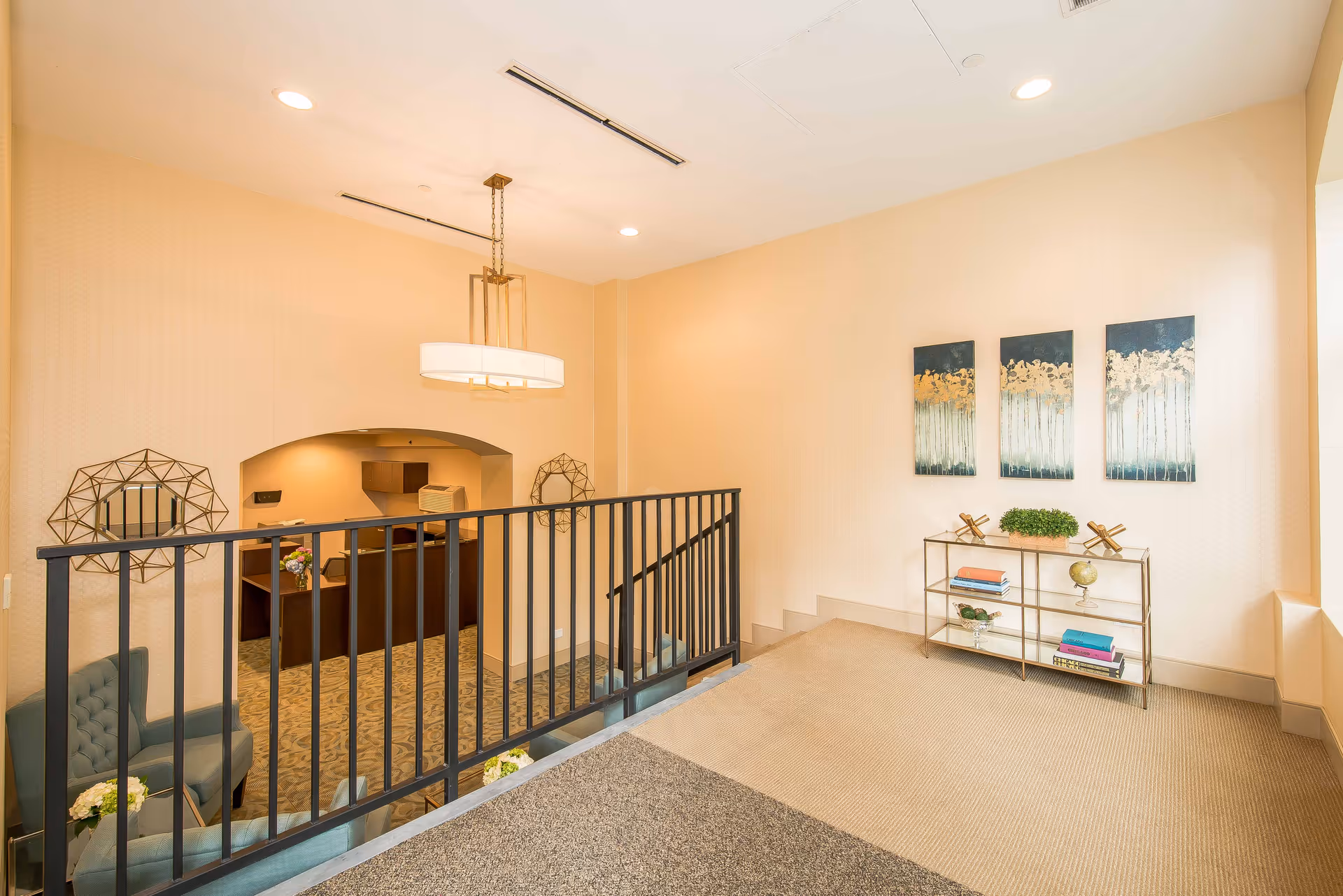 Interior view of a senior living facility featuring a carpeted landing area with a metal railing overlooking a reception desk below. The landing area has a small decorative shelf with books, plants, and ornaments, and three abstract paintings on the wall. There are two blue armchairs with a small table and flowers near the railing. The space is well-lit with ceiling lights and a hanging light fixture.