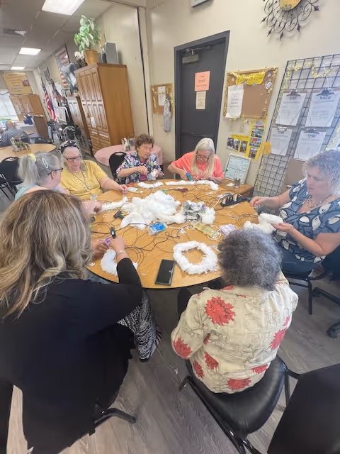A group of elderly women sitting around a large round table engaged in a craft activity involving white materials, in a well-lit room with bulletin boards and cabinets in the background.