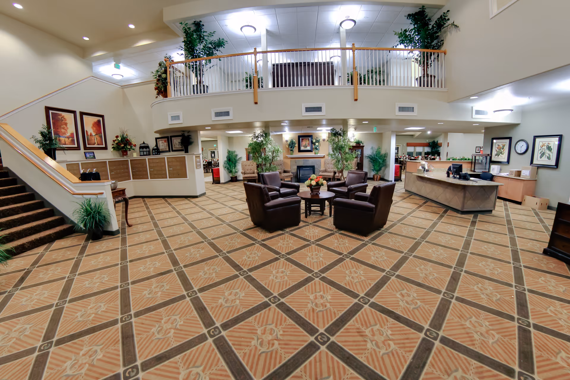 Spacious two-story senior living lobby with a central seating area, reception desk, staircase and decorative plants.