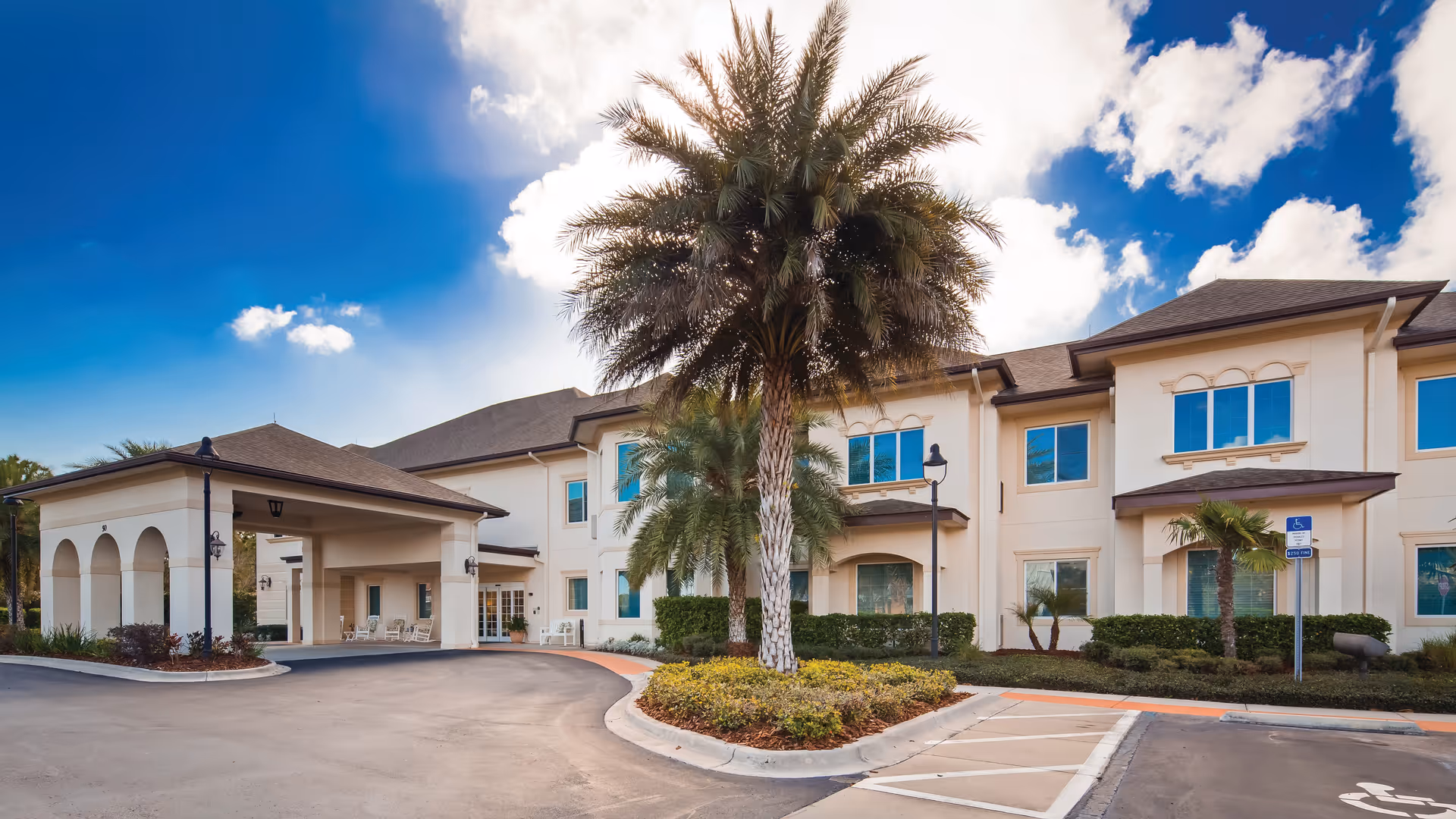 Exterior view of The Windsor of Palm Coast senior living facility showing a two-story building with beige walls, multiple windows, a covered entrance with arches, palm trees, and a clear blue sky with some clouds.