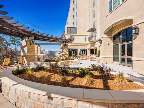 Sunny outdoor courtyard with a curved raised stone planter, pergola, seating, and the building's entrance under a blue sky.