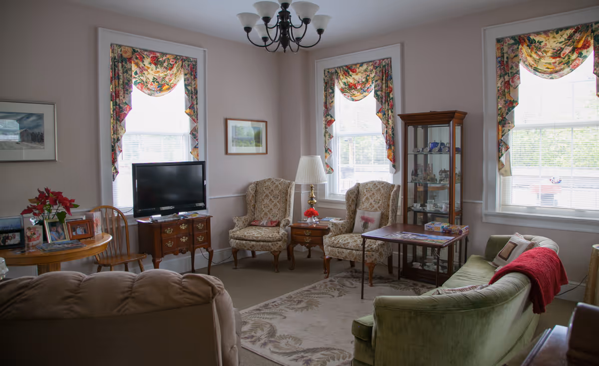 A cozy living room with floral curtains on three windows, two patterned armchairs, a green sofa with a red throw blanket, a wooden display cabinet with glass shelves, a TV on a wooden stand, a small table with a puzzle, and a round table with framed photos and a poinsettia plant.