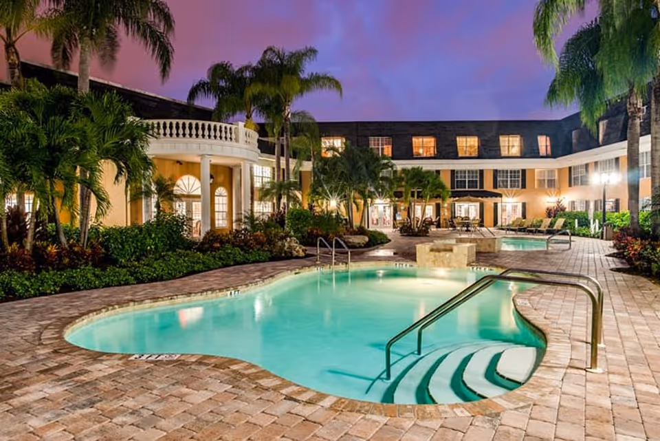 Outdoor swimming pool area at dusk with palm trees and a multi-story building in the background, featuring warm lighting from windows and patio furniture around the pool.