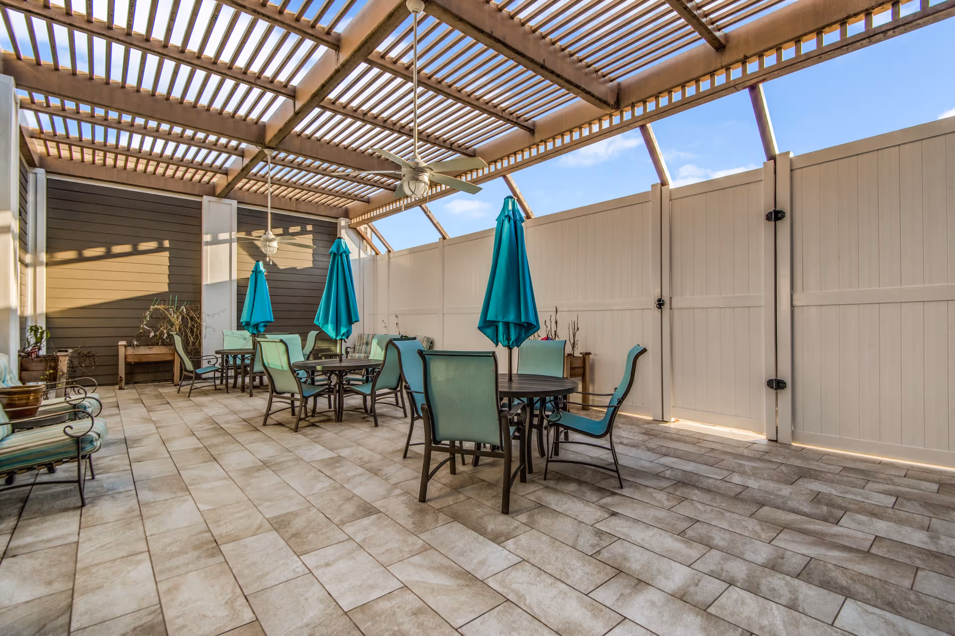 Covered outdoor patio area with tiled floor, several round tables with teal umbrellas and matching chairs, ceiling fans, and a white privacy fence under a slatted pergola roof.