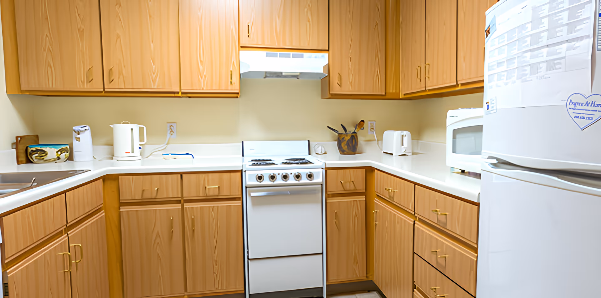 A compact kitchen with light wood cabinets and white countertops. The kitchen features a white stove with four burners, a white refrigerator with papers and a heart-shaped note attached, a microwave, a toaster, an electric kettle, and a utensil holder with wooden spoons. The walls are painted a light color and there is a range hood above the stove.
