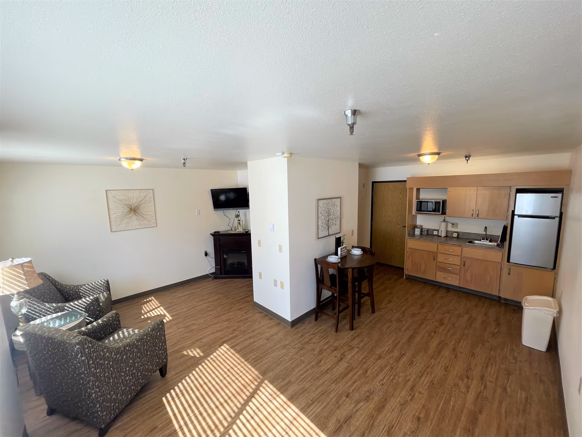 A bright and spacious senior living apartment featuring a small kitchenette with wooden cabinets, a microwave, and a refrigerator. There is a small round dining table with two chairs set with bowls and plates. The living area has two patterned armchairs and a side table with a lamp. A flat-screen TV is mounted on the wall above a small electric fireplace. The room has wood-style flooring and white walls with minimal artwork.