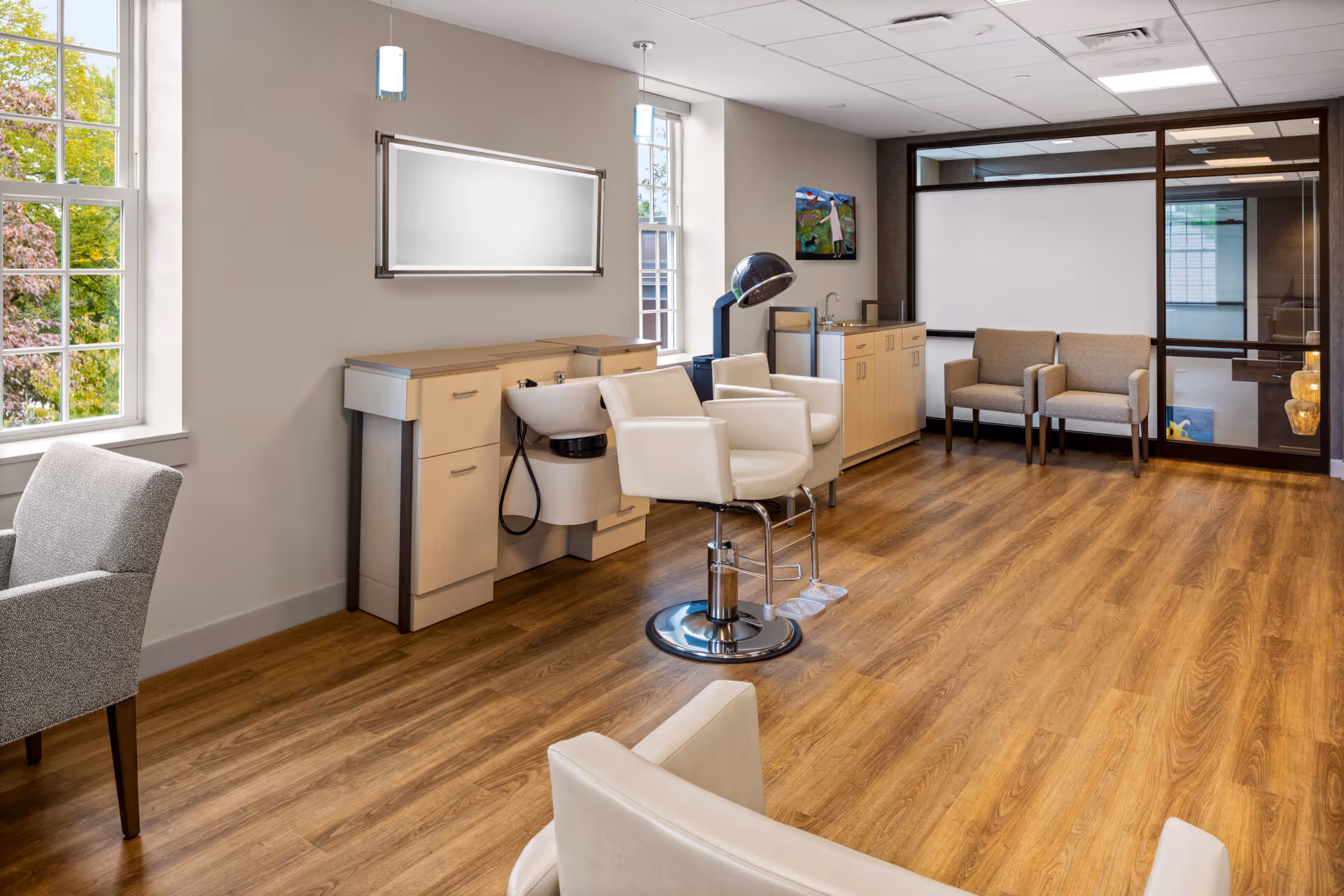 A bright and clean hair salon area inside a senior living facility with wooden flooring, beige walls, and large windows letting in natural light. The room features a salon chair in front of a hair washing station with cabinets, a hair dryer hood, and additional seating with armchairs along the walls. A colorful painting hangs on the wall near the back, and a large glass partition separates this space from another room.