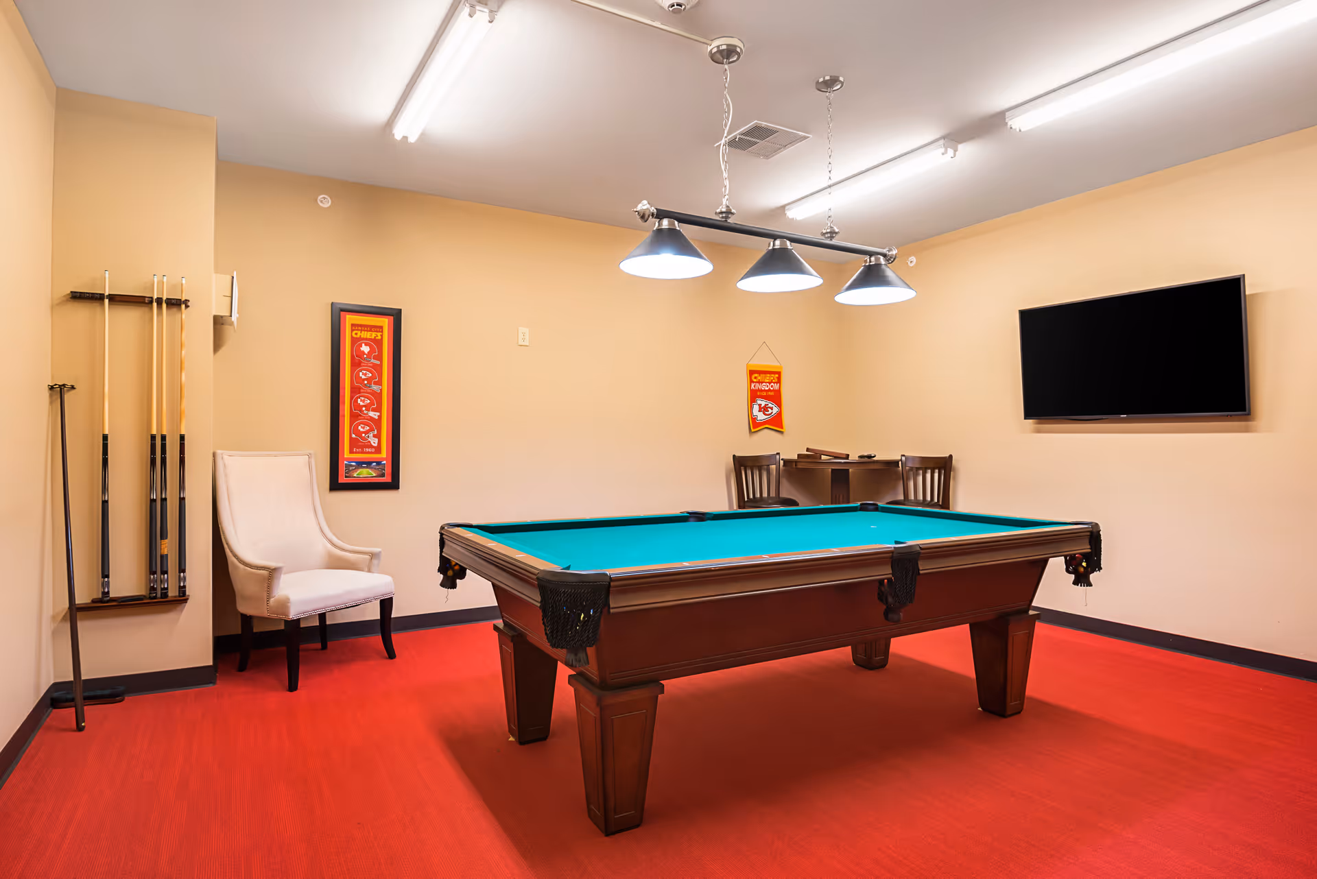 A game room with a pool table in the center, a rack with pool cues on the left wall, a beige upholstered chair, a small table with two chairs in the back corner, a flat-screen TV mounted on the right wall, and Kansas City Chiefs decorations on the walls. The room has red carpet and beige walls with overhead lighting.