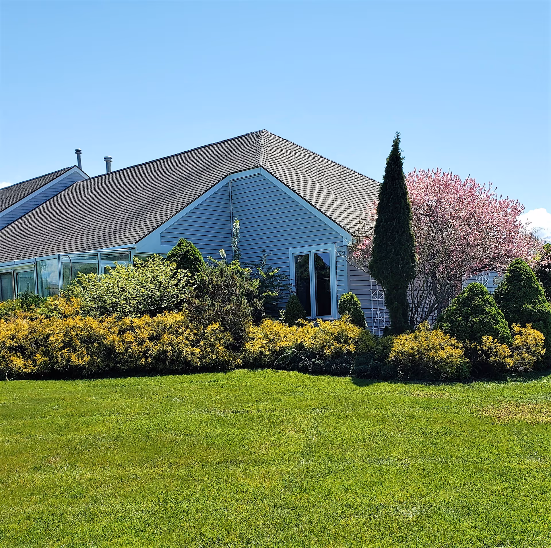 Exterior view of a single-story building with a sloped roof, surrounded by well-maintained green grass, various shrubs, and a blooming pink tree under a clear blue sky.