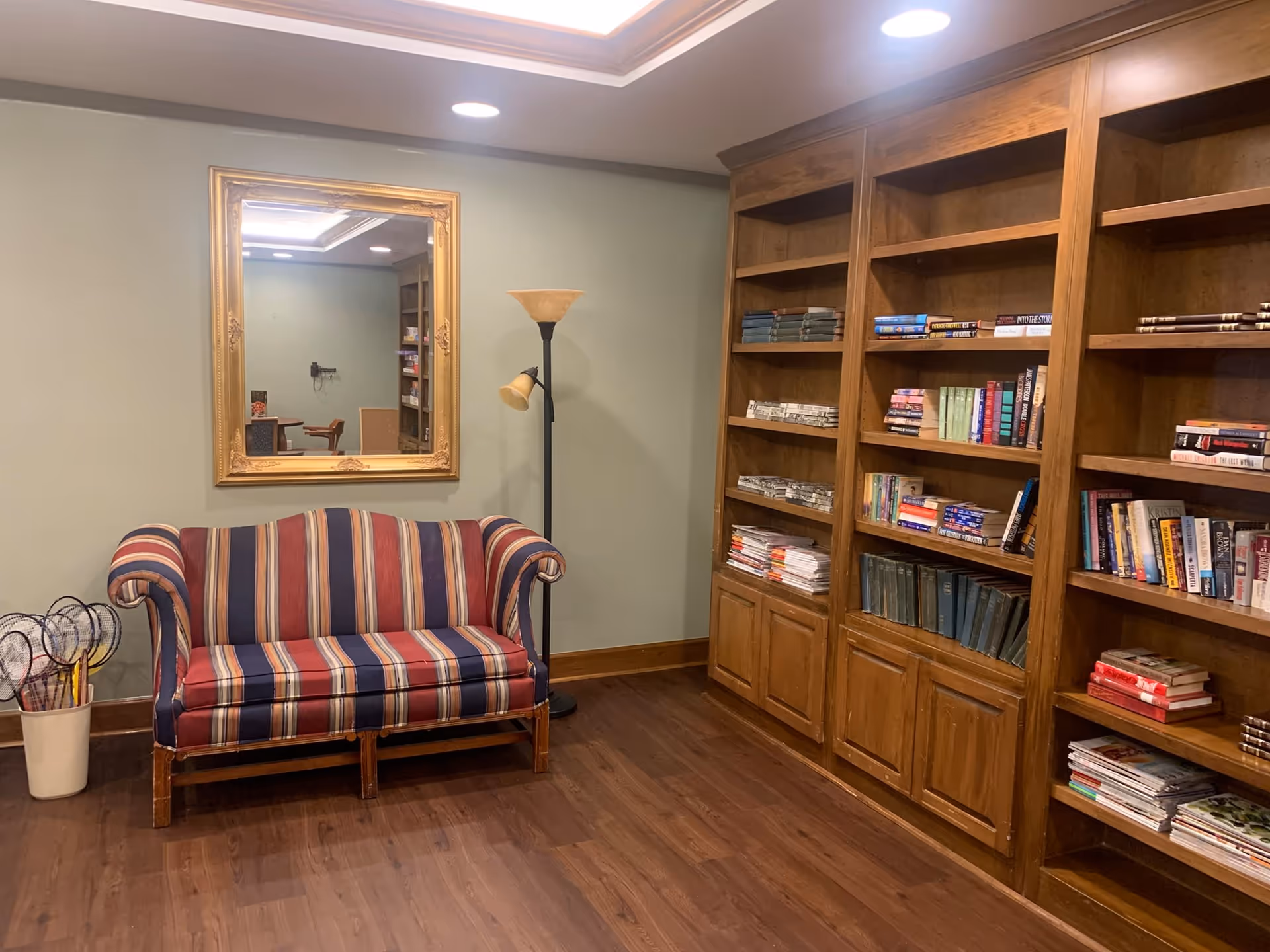 A cozy room with a striped upholstered loveseat, a tall floor lamp, a large gold-framed mirror on the wall, and wooden bookshelves filled with books and magazines. The room has wooden flooring and soft lighting.