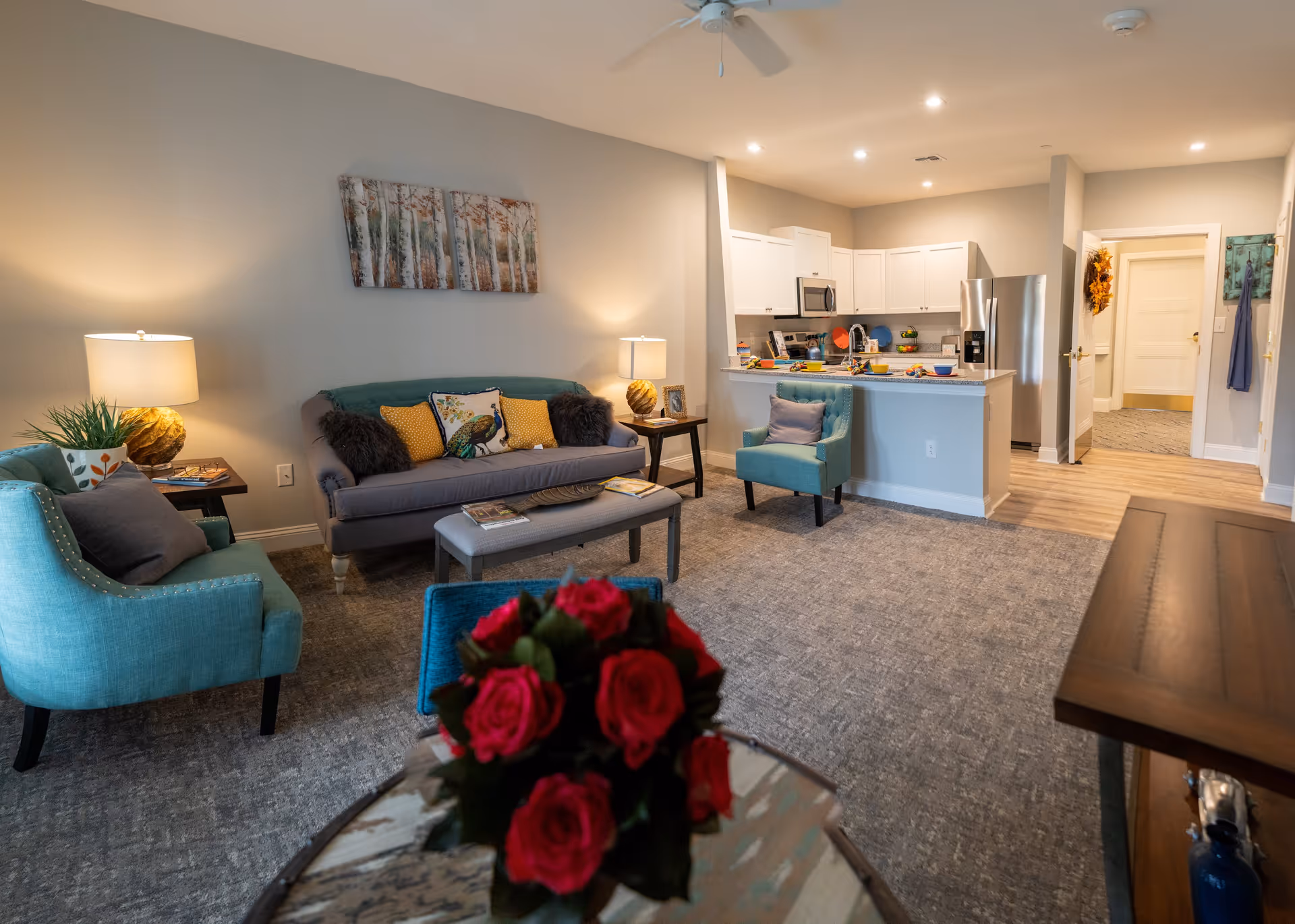 A cozy living room area with a gray sofa adorned with colorful pillows, two teal armchairs, a small ottoman, and side tables with lamps. The room opens into a kitchen with white cabinets, stainless steel appliances, and a breakfast bar. A bouquet of red roses is in the foreground on a glass table.