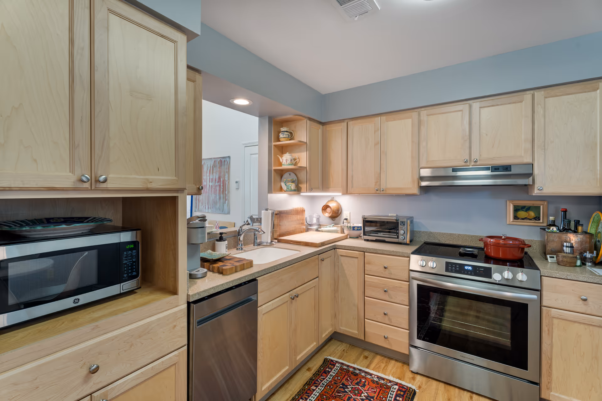 A modern kitchen with light wood cabinets, stainless steel appliances including a microwave, dishwasher, and oven with a red pot on the stove. The kitchen features a double sink, a small toaster oven, and various kitchen utensils and decor items on the countertops. There is a colorful rug on the wooden floor.