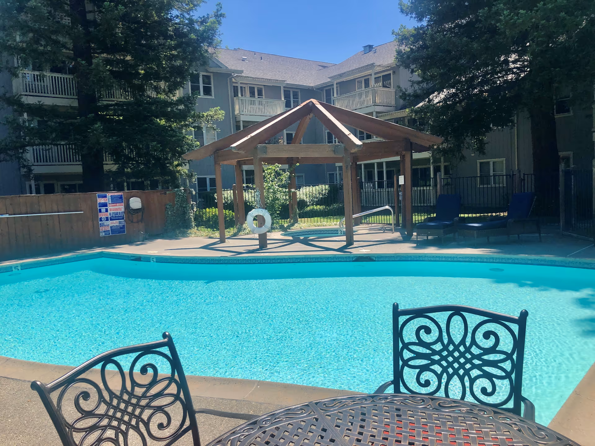 Outdoor swimming pool area with clear blue water, surrounded by a concrete deck. In the foreground, there is a decorative metal table and chairs. In the background, there is a wooden pergola structure with a life preserver hanging from it, two blue lounge chairs, and a multi-story residential building with balconies. Trees and greenery surround the pool area under a clear blue sky.