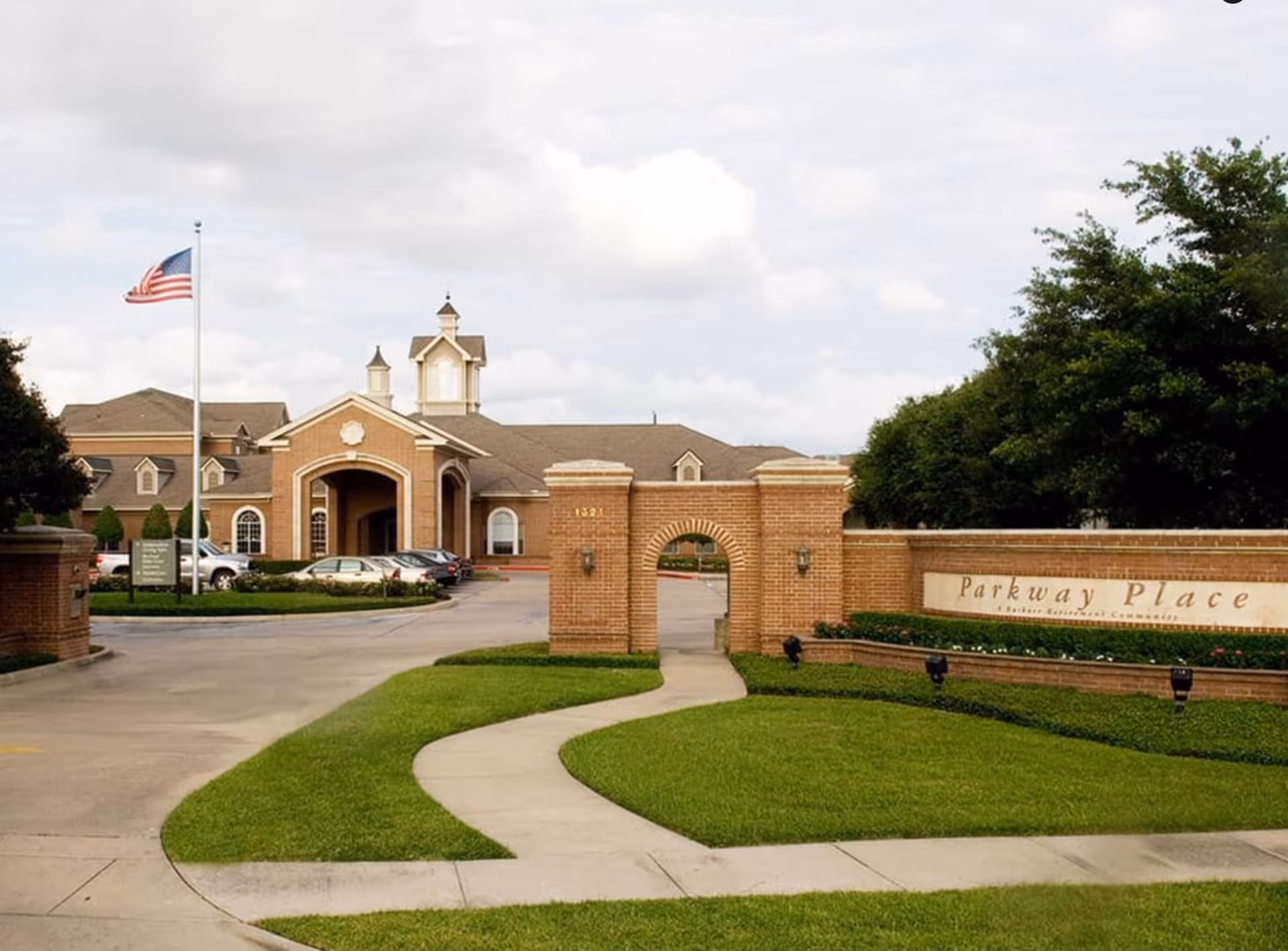 Exterior view of Buckner Parkway Place senior living facility showing a brick entrance with an archway, a curved sidewalk, well-maintained green lawn, an American flag on a flagpole, and the main building with a peaked roof and cupolas in the background under a cloudy sky.