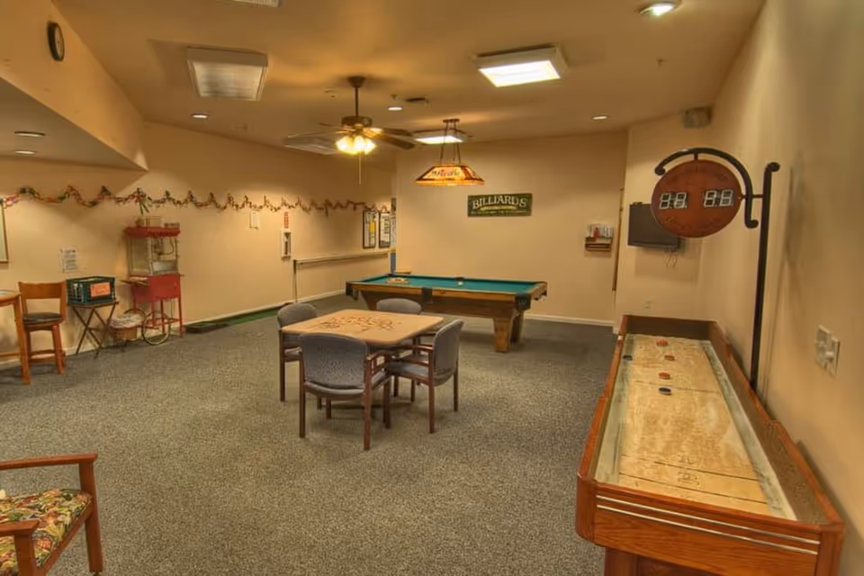 A recreational room with a pool table, a shuffleboard table, a card table with four chairs, a popcorn machine, and additional seating. The walls are decorated with a garland and a sign that says 'Billiards'. The room has carpeted flooring and ceiling lights including a ceiling fan.