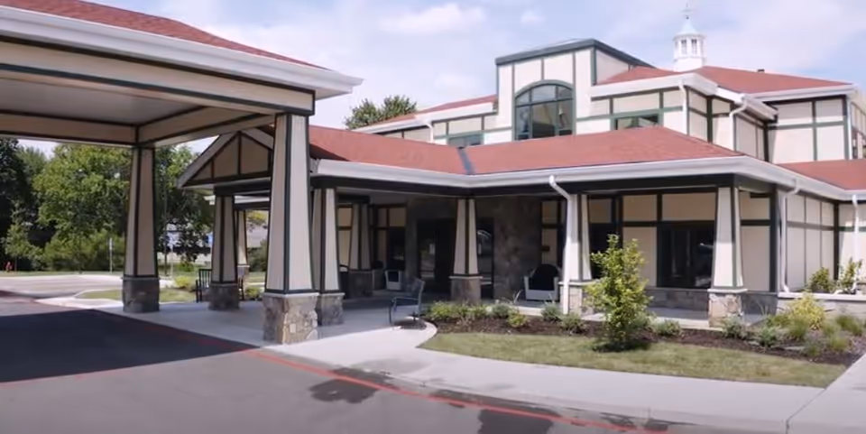 Exterior view of Creekside at Elfindale Assisted Living building entrance with covered drop-off area, stone pillars, and landscaped greenery under a partly cloudy sky.