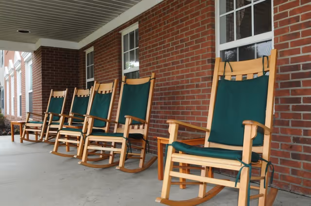 A row of wooden rocking chairs with green cushions lined up on a covered porch with a brick wall and windows in the background.