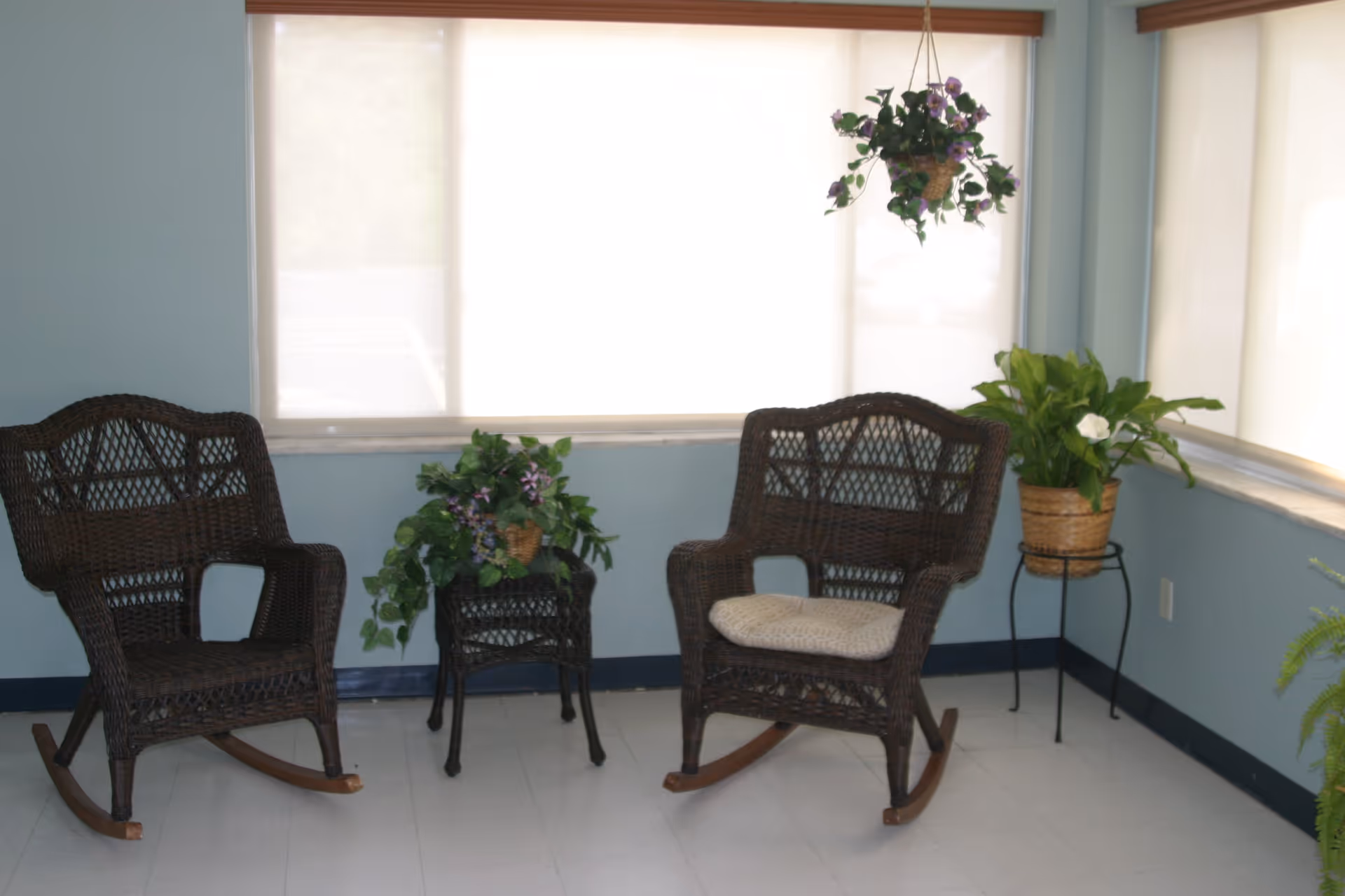 A small seating area with two dark wicker rocking chairs, one with a cushion, placed on a light-colored floor in front of windows with white blinds. There are three potted plants: one on a small wicker table between the chairs, one hanging from the ceiling, and one on a metal plant stand in the corner.