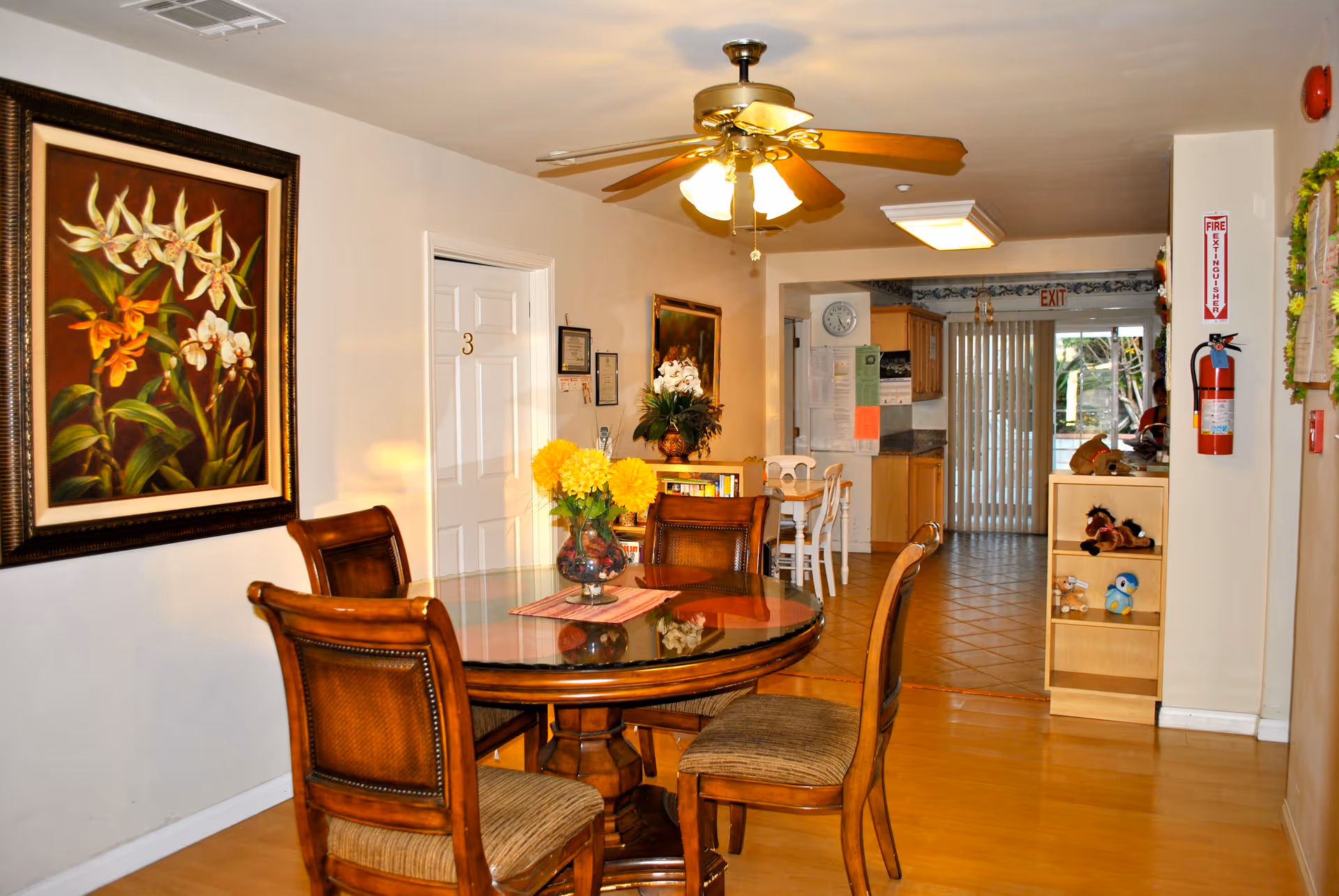 Interior view of a senior living facility dining area with a round wooden table and four chairs. A vase with yellow flowers is on the table. The room has a ceiling fan with lights, a large floral painting on the wall, and a door labeled with the number 3. In the background, there is a kitchen area with wooden cabinets, a clock on the wall, and sliding glass doors leading outside. A fire extinguisher is mounted on the wall near a small shelf with stuffed animals.