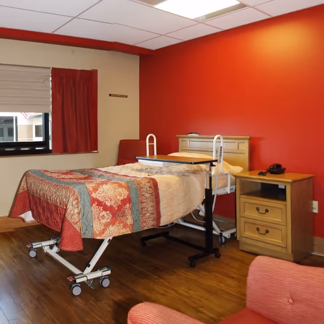A bedroom in an assisted living facility featuring a hospital-style bed with a patterned quilt, a wooden nightstand with a telephone, a red accent wall, a window with beige and red curtains, and a red upholstered chair in the foreground.