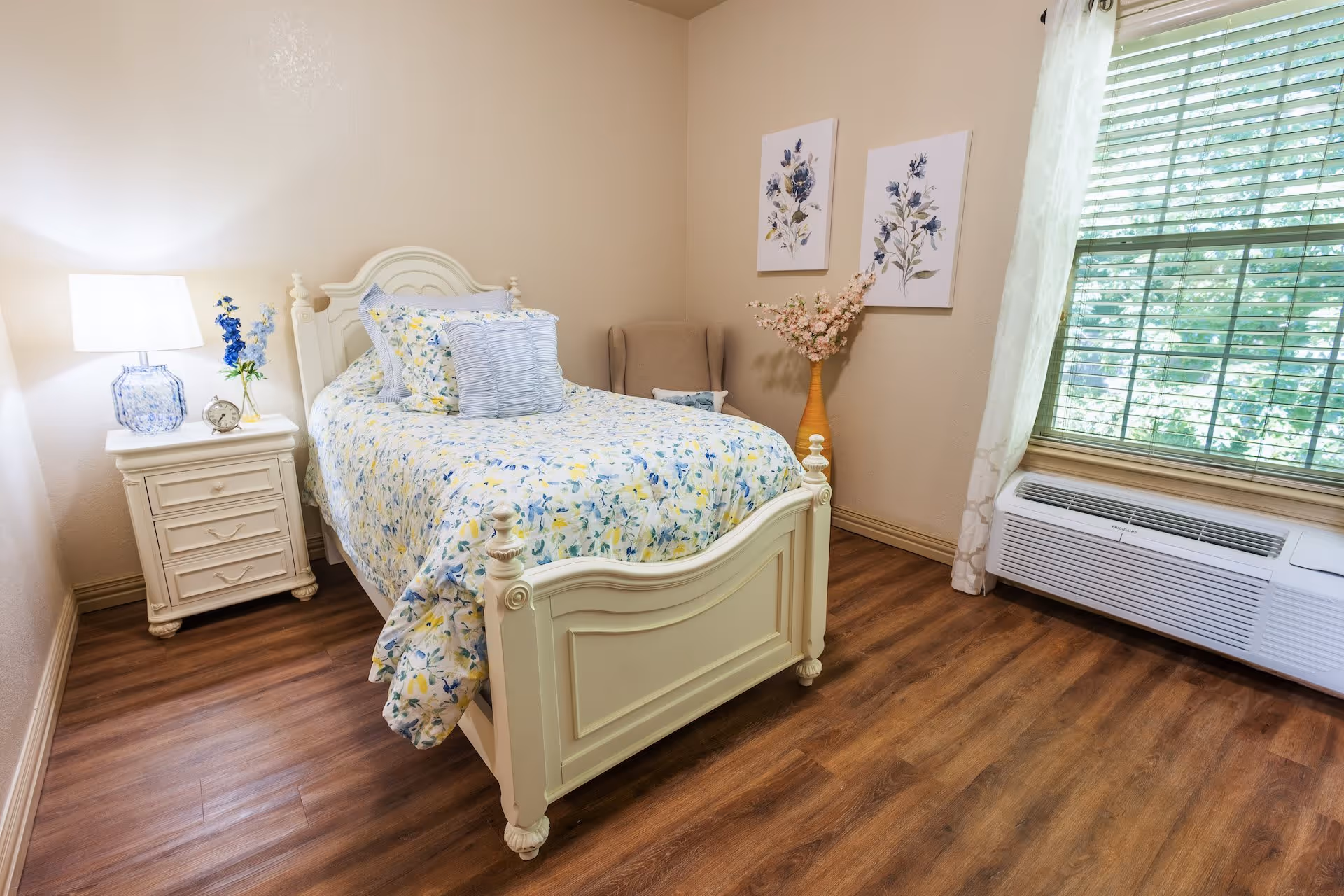 A cozy bedroom with a white wooden bed featuring floral bedding in shades of blue and yellow. Next to the bed is a white nightstand with a blue and white lamp, a small clock, and a vase with blue flowers. The room has wooden flooring, a window with white curtains and blinds, and two floral paintings on the wall above a beige armchair with a vase of pink flowers beside it.