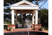 A white gazebo with a peaked roof and four columns, surrounded by trees and greenery. In front of the gazebo is a circular water fountain with water spraying upwards. There are potted plants on either side of the brick base of the gazebo.