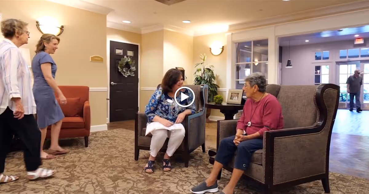 A cozy seating area in a senior living facility with two women sitting and conversing on armchairs. Two other women are walking past, and a man is visible in the background near the entrance. The room has warm lighting, a patterned carpet, and neutral-colored walls with a door decorated with a wreath.