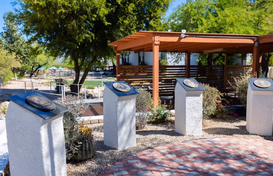 Outdoor garden area with four white stone pedestals topped with plaques, surrounded by gravel and plants. A wooden pergola structure is visible in the background along with trees and a paved walkway.