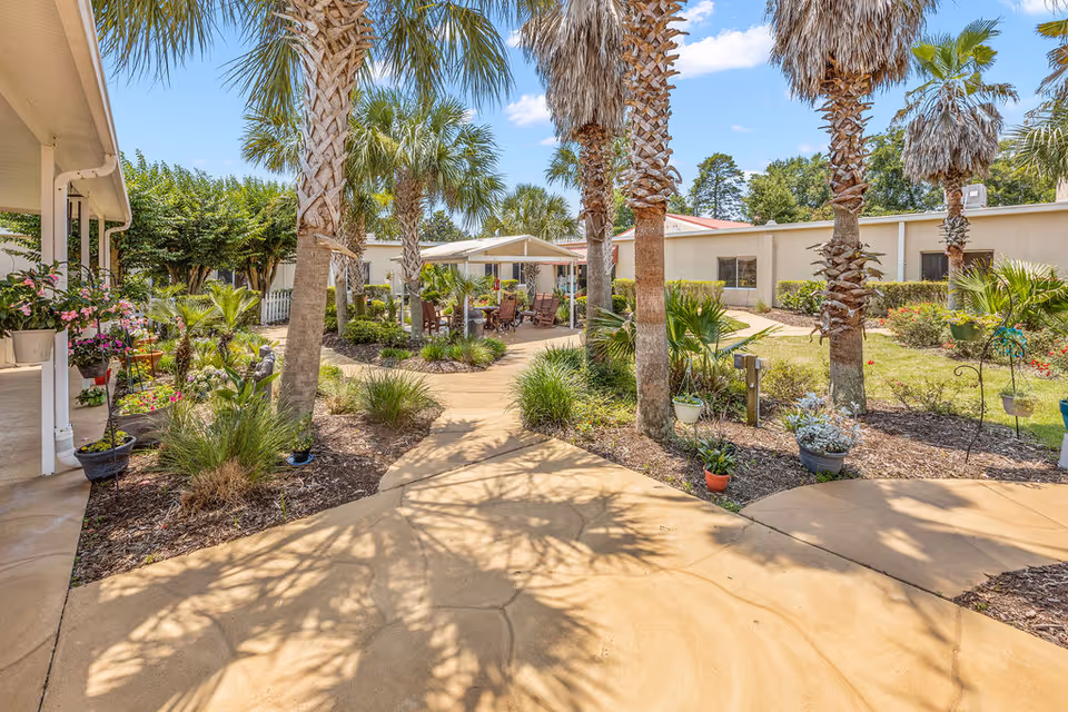 Outdoor courtyard area at Manor at Bluewater Bay featuring a paved walkway surrounded by palm trees, various plants, and flowers. There is a covered seating area with chairs and tables in the background, and the building exterior is visible under a clear blue sky.