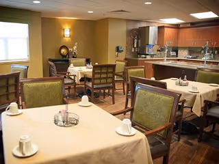 A dining room in a senior living facility with several tables covered in beige tablecloths, each set with white cups and saucers. The room has wooden chairs with green cushions, a window letting in natural light, and a kitchen area with cabinets and countertops in the background.