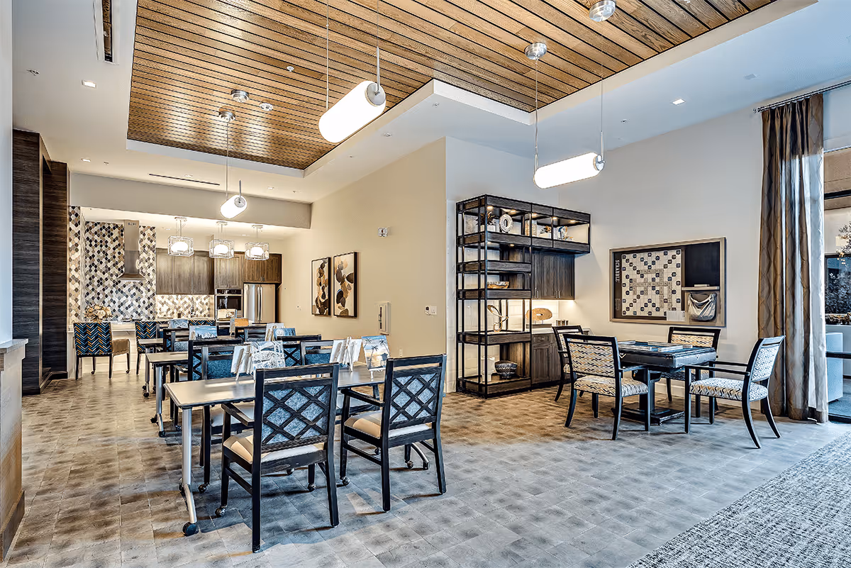 A spacious, modern dining area with multiple tables and chairs arranged neatly. The room features a wooden ceiling with hanging pendant lights, a kitchen area with a tiled backsplash and stainless steel appliances in the background, and a shelving unit with decorative items. There is also a game board mounted on the wall near a set of chairs and a table, with large windows covered by curtains letting in natural light.
