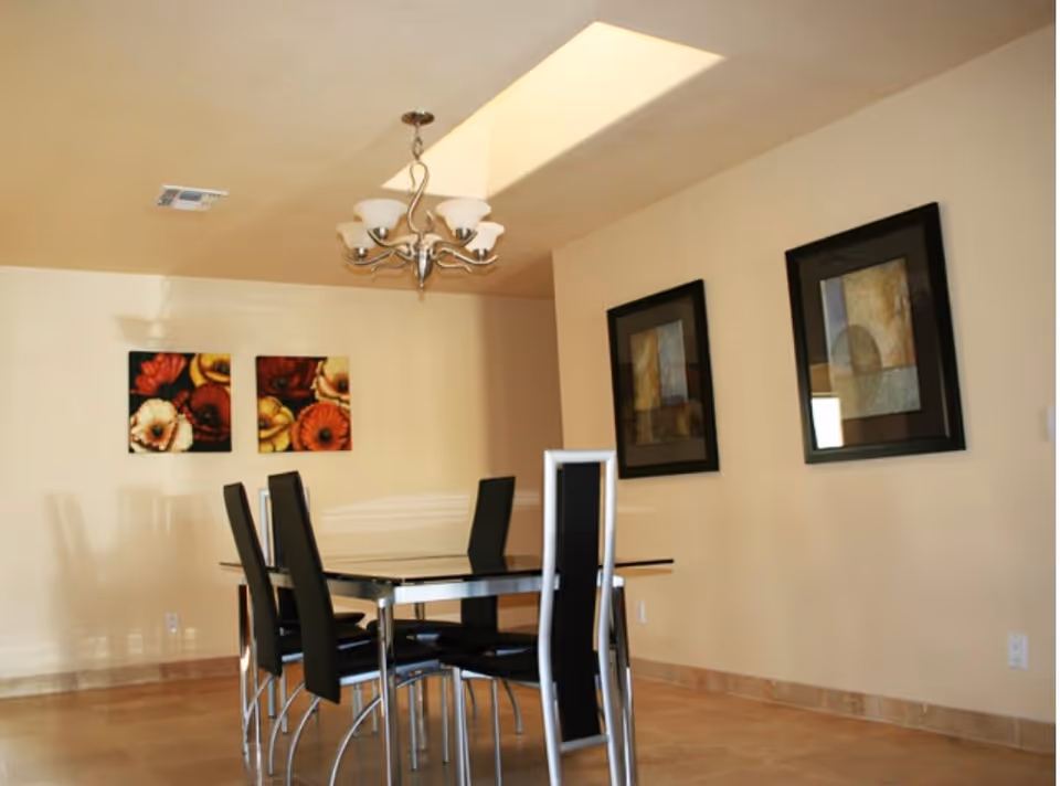 A dining room with a rectangular glass table and six black high-back chairs. The walls are beige with two floral paintings on one wall and two abstract framed artworks on another. A modern chandelier hangs from the ceiling, and the floor is tiled.
