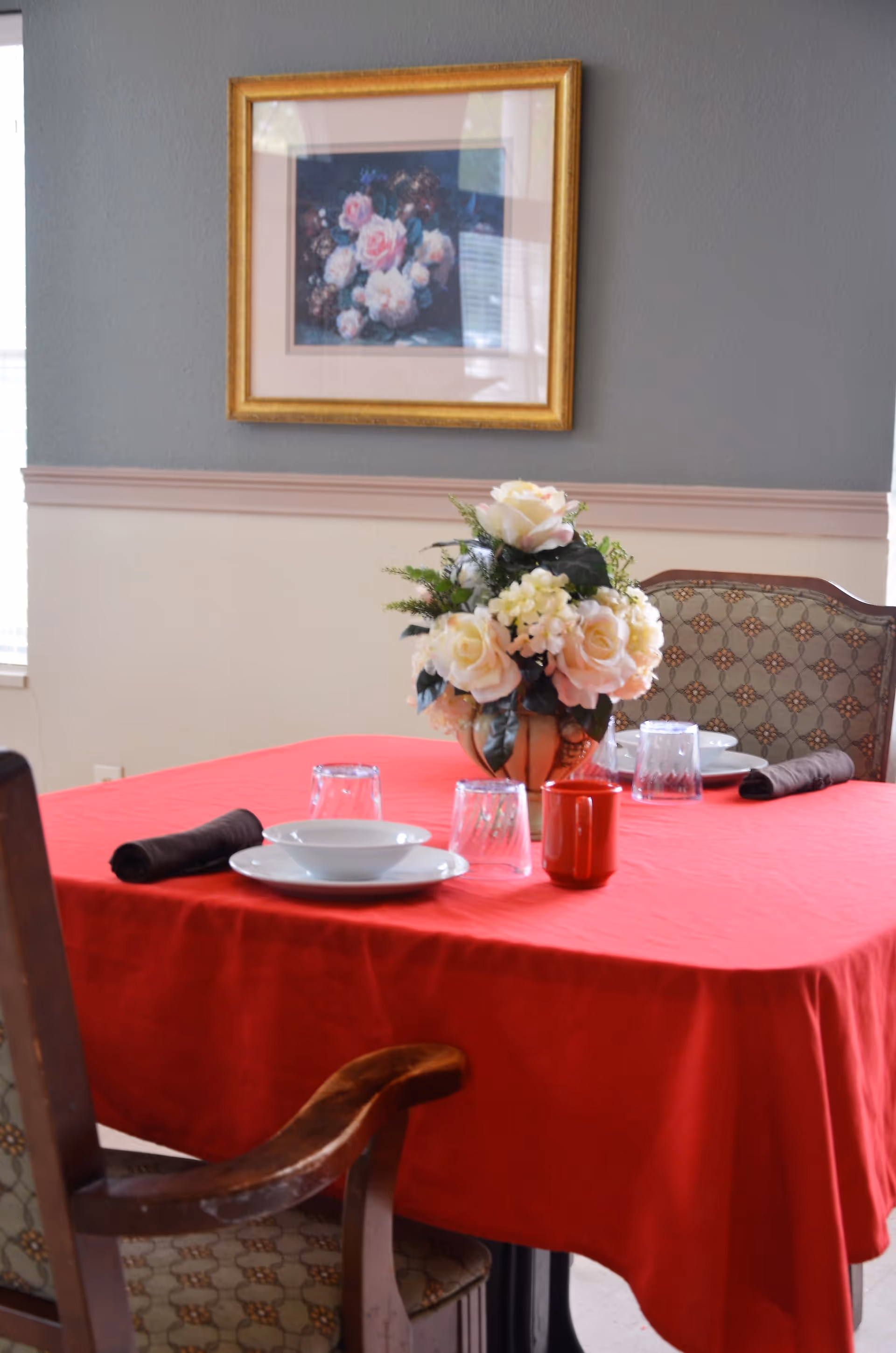A dining table covered with a red tablecloth set with white plates, upside-down clear glasses, dark cloth napkins, and a red mug. A floral centerpiece with white and pale pink flowers is placed in the middle of the table. Two upholstered chairs with patterned fabric are positioned around the table. A framed painting of flowers hangs on the gray and beige wall behind the table.