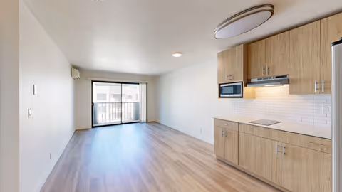 Open-plan living area with wood-look flooring, a kitchenette on the right, and a sliding glass door to a balcony.