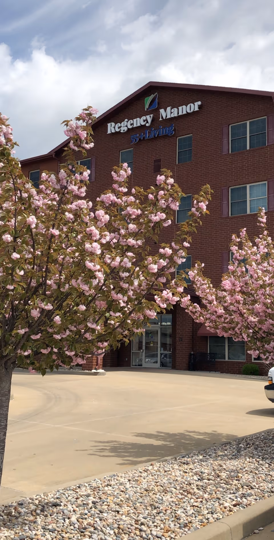Exterior view of Regency Manor 55+ Independent Senior Living Apartments building with blooming pink flowering trees in the foreground and a clear sky above.