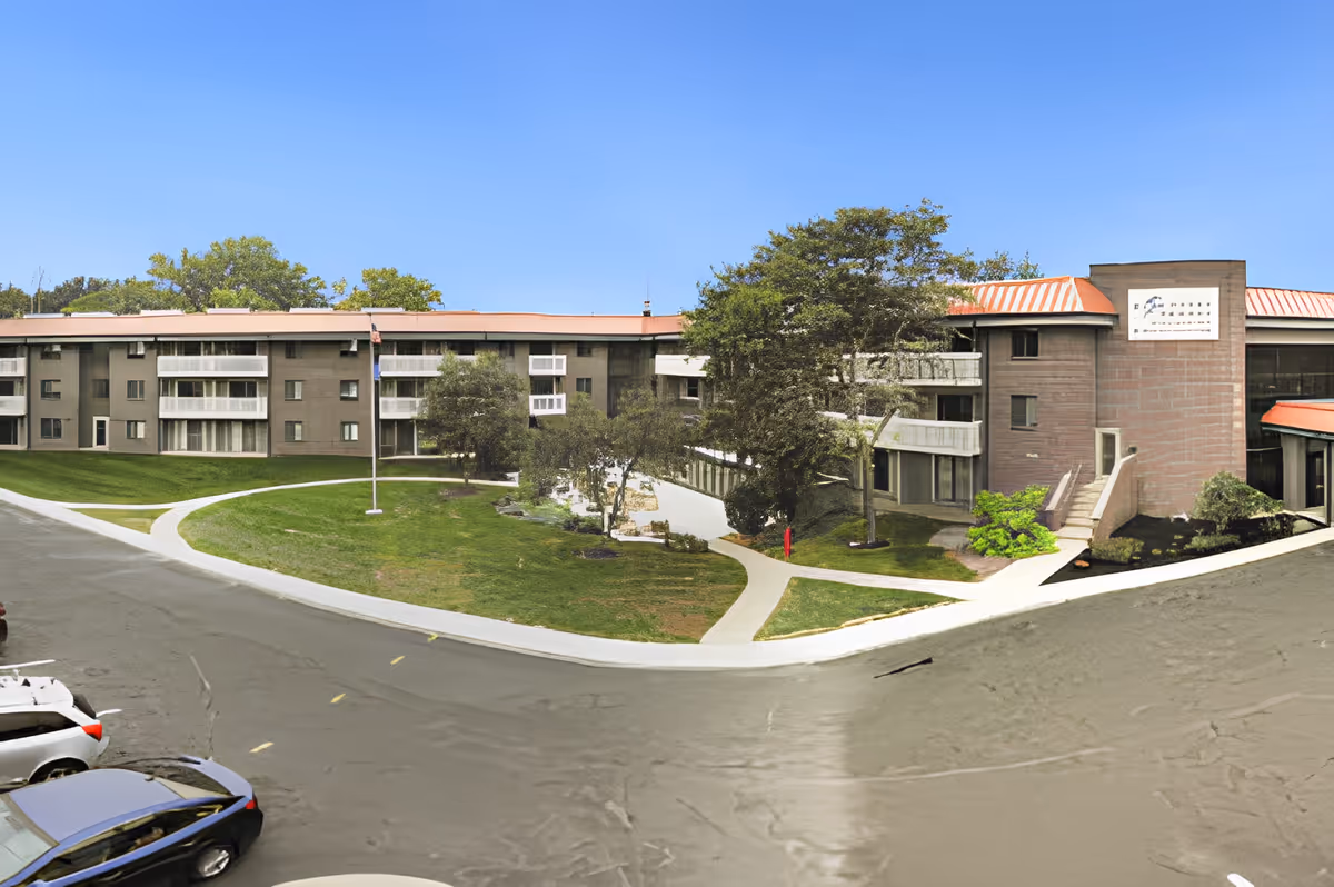 Front exterior of a three-story senior living building with balconies, landscaped lawn and walkways, and parked cars.