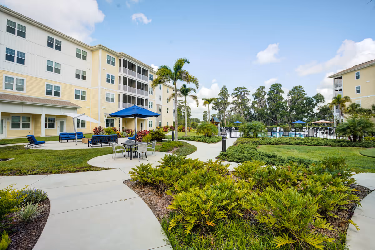 Outdoor courtyard area of a senior living facility with paved walkways, green landscaping, palm trees, and patio furniture including tables with umbrellas and lounge chairs. The building is a multi-story structure painted in yellow and white with multiple windows and screened balconies. A fenced swimming pool area is visible in the background under a partly cloudy sky.
