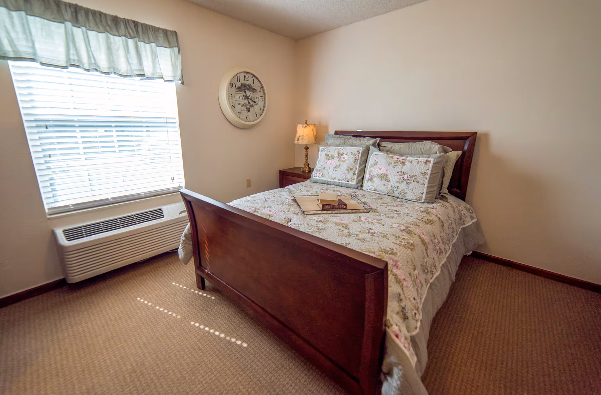 A cozy bedroom with a wooden bed frame and floral bedding. There are two matching floral pillows and a tray with books on the bed. A bedside table with a decorative lamp is next to the bed. A large window with blinds and a valance lets in natural light. A round wall clock hangs on the wall above the bedside table.