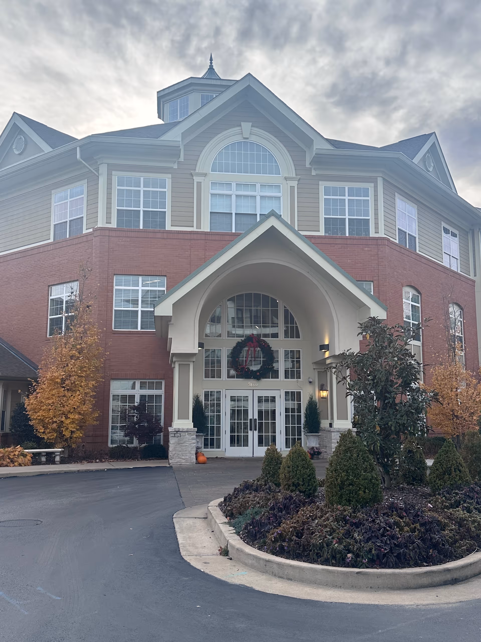 Front exterior view of a multi-story assisted living and memory care facility with a large arched entrance, decorated with a holiday wreath above the double glass doors. The building has a combination of brick and siding, multiple windows, and a landscaped circular driveway with shrubs and small trees.