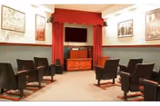 A small theater room with rows of black cushioned chairs facing a large flat-screen TV mounted on a wooden cabinet. The walls are decorated with framed pictures and the TV area is framed with red curtains and a red valance.
