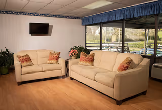 A cozy living room area with two beige sofas adorned with patterned throw pillows. A flat-screen TV is mounted on the wall above one sofa. There are potted plants and a side table with a flower arrangement between the sofas. Large windows with blue valances provide a view of an outdoor patio and greenery.