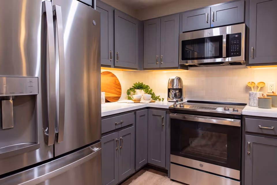 Modern kitchen with stainless steel refrigerator, oven, and microwave. Gray cabinets with silver handles, white countertops, a coffee maker, wooden utensils in a holder, and a small green plant on the counter.
